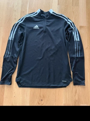 Svart långärmad träningströja Adidas - Snygg svart långärmad träningströja från Adidas med vita ränder på ärmarna och dragkedja vid halsen. Tillverkad i ett tekniskt funktionsmaterial med Aeroready-tryck. Perfekt för träning eller uppvärmning.