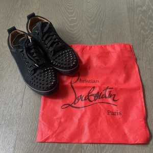 Svarta sneakers från Christian Louboutin - Snygga svarta sneakers från Christian Louboutin med ikoniska röda sulor. Skorna har snörning och är dekorerade med nitar på tån för en edgy look. Perfekta för att ge din outfit en lyxig touch.