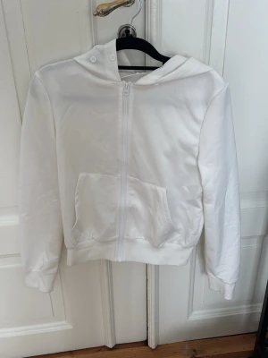 Vit hoodie med dragkedja - Säljer en stilren vit hoodie med dragkedja framtill och stor ficka på magen. Hoodien har huva och ribbade muddar vid ärmslut och nederkant. Perfekt för dig som gillar en enkel och clean look. Materialet känns mjukt och bekvämt.