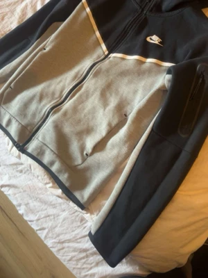 Nike hoodie i grått och svart - Snygg hoodie från Nike i grått och svart med vit logga på bröstet. Dragkedja framtill, två fickor och en extra ficka med dragkedja på ärmen. Stilsäker design med vita detaljer och huva. Perfekt för en sportig och avslappnad look.