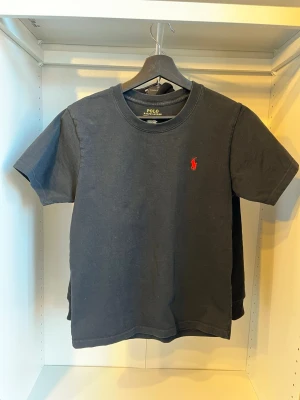 Ralph Lauren T-shirt  - Säljer en väldigt snygg Ralph Lauren t-shirt. Svart med ett rött polo märke. Du får även en hållare för tröjan. Inga hål eller defekter. Storlek: xs ( 170-173cm )