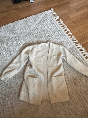 Beige ribbad kofta från H&M - Säljer en beige, lång och öppen kofta från Gina Tricot. Koftan har ribbad struktur, långa ärmar och två fickor framtill. Perfekt att slänga över en t-shirt för en chill look.