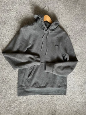 Mörkgrå hoodie från J.Lindeberg - Mörkgrå hoodie från J.Lindeberg med klassisk passform, ribbade muddar och en stor ficka framtill. Materialet är mjukt och bekvämt, perfekt för chill dagar. Diskret logga på bröstet och justerbar huva med snören.