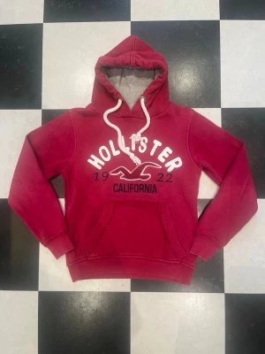 Röd hoodie från Hollister - Cool röd hoodie från Hollister med vit broderad logga och text 'California' på bröstet. Den har en stor huva med vita snören och en klassisk magficka. Perfekt för dig som gillar avslappnad stil och vill ha något mjukt och bekvämt.
