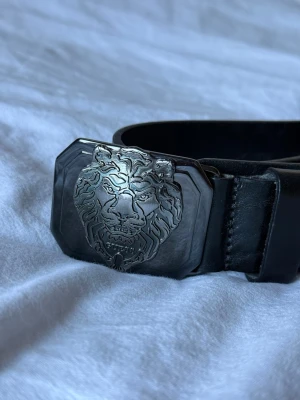 Svart Guess bälte  - Snyggt svart bälte från Guess med ett stort, silverfärgat spänne prytt av ett detaljerat lejonhuvud. Bältet har präglad logga längs remmen och är gjort i skinn. Perfekt för att lyfta din outfit med en cool detalj. Notera bältet har några hål i sig som gjorts i efterhand för egen justering av storlek. 