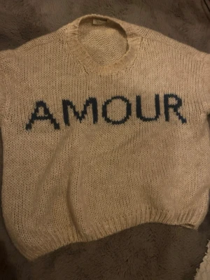 Beige stickad tröja med text - Stickad beige tröja med rund halsringning och svart texten 'AMOUR' framtill. Tröjan har en avslappnad passform och ribbstickade muddar vid hals och nederkant. Perfekt för en trendig och mysig look.