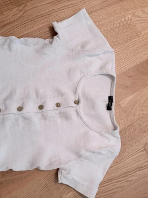 Vit ribbad topp med knappar  - Vit ribbad top, korta ärmar och rund halsringning. Toppen har dekorativa knappar framtill och är tillverkad i ett mjukt, stretchigt material som sitter snyggt på kroppen.