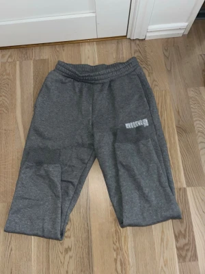 Gråa mjukisbyxor från Puma - Snygga grå mjukisbyxor från Puma med tryckt logga på ena benet. Byxorna har resår i midjan och är tillverkade i mjukt material som känns skönt mot huden. Perfekta för en avslappnad stil och passar till hoodie eller t-shirt.