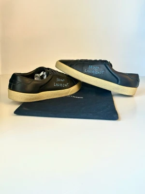 Svarta Saint Laurent sneakers i läder - Svarta sneakers från Saint Laurent i mjukt läder med handskriven logga på sidan. Skorna har en klassisk låg profil och beige gummisula som ger en clean och stilren look. Perfekta för dig som gillar exklusiva och minimalistiska sneakers.