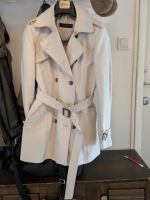 Beige trenchcoat från Zara Woman - Stilren beige trenchcoat från Zara Woman med klassisk dubbelknäppning, bälte i midjan och snygga axelklaffar. Jackan har fickor framtill och spänne vid ärmsluten. Perfekt för dig som vill ha en tidlös och trendig look.