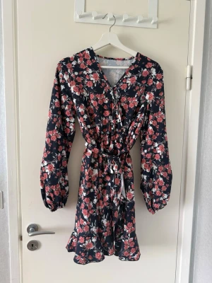 Klänning - Supersöt kort omlottklänning med blommigt mönster i rött, vitt och rosa på mörkblå botten. Klänningen har långa ärmar, v-ringning, volangkant nertill och knyt i midjan för en snygg passform. Perfekt för sommarens alla tillfällen!