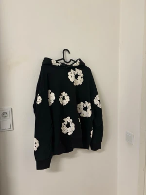 Svart hoodie med vita blommor - Cool svart hoodie med stora vita blommor tryckta över hela tröjan. Hoodien har huva och en klassisk känguruficka framtill. Perfekt för dig som gillar streetwear och vill sticka ut med ett unikt mönster. Mjuk och skön bomullskänsla.