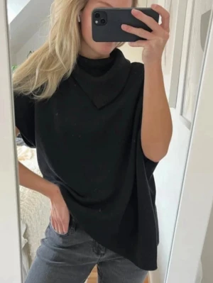 Svart ponchotröja med hög krage - Snygg svart ponchotröja med lös passform och hög krage. Perfekt för lager-på-lager och ger en chill vibe till din outfit. Tröjan har korta ärmar och är tillverkad i ett mjukt material som faller snyggt.