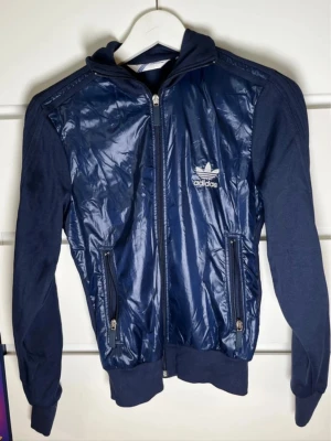 Blå vindjacka från Adidas - Snygg marinblå vindjacka från Adidas med glansig framsida och klassisk logga på bröstet. Jackan har dragkedja framtill, två fickor med dragkedja och ribbade muddar. Sportig. 