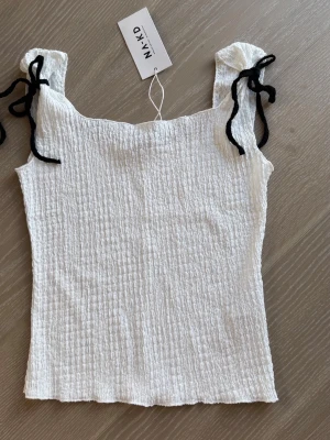 Vit linnetopp från NA-KD i stl S helt ny - Superfin vit linnetopp från NA-KD med smockat tyg och svarta rosetter på axlarna. Toppen är ärmlös och har en fyrkantig ringning. Helt ny 