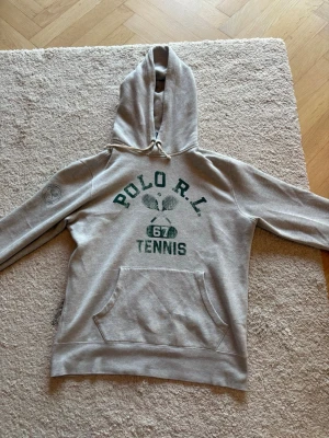 Grå hoodie Polo Ralph Lauren Tennis - Grå hoodie från Polo Ralph Lauren med grön text och tennistryck på bröstet. Stor känguruficka framtill och dragsko i huvan. Mjuk insida och broderad detalj på ärmen. Perfekt för en sportig och avslappnad stil.