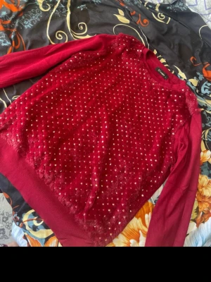 Röd tröja med paljetter från Vero Moda - Röd tröja  med fluffig framsida och massor av små guldiga paljetter som ger ett glittrigt intryck. Tröjan har rund hals och långa ärmar i stickat material, perfekt för att sticka ut i mängden.