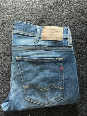 Blå Replay jeans med smal passform - Säljer ett par klassiska blå jeans från Replay med snygga detaljer på bakfickan och Replay-lapp i midjan. Och en liten slitning på höger knä för lite mer detaljer på dem. Jeansen har go passform och är tillverkade i mjuk bomull. Perfekta för dig som gillar en stilren och avslappnad look. 