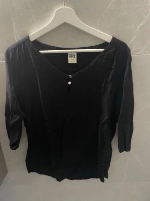 Svart blus med knappar Vero Moda - Stilren svart blus från Vero Moda med v-ringning och två dekorativa knappar framtill. Blusen har trekvartsärm och är tillverkad i ett lätt, mjukt tyg.