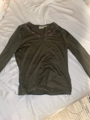 Olivgrön topp med spets från Gina Tricot - Snygg olivgrön långärmad topp från Gina Tricot med v-ringning och dekorativ spetsdetalj runt halsen. Perfekt för dig som gillar en enkel men ändå stylish look. Materialet känns mjukt och bekvämt mot huden.