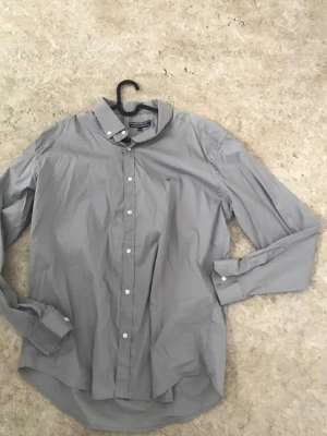 Grå skjorta från Tommy Hilfiger - Snygg grå skjorta från Tommy Hilfiger med klassisk krage och knappar framtill. Skjortan har lång ärm och en liten broderad logga på bröstet. Perfekt till jeans eller chinos för en clean look.