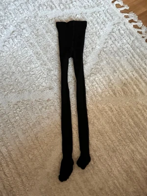 Svarta tights i klassisk modell - Svarta tights med hög midja och helfot, perfekta att bära under kjol eller klänning. Tightsen är stretchiga och sitter tajt längs benen. Materialet är mjukt och följsamt, vilket gör dem bekväma hela dagen.