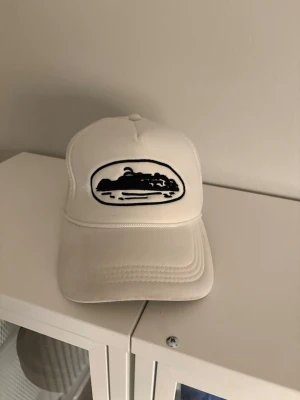 Vit meshkeps med svart broderi - Vit keps med mesh på sidorna och bak, samt ett svart broderat motiv på fronten. Kepsen har böjd skärm och justerbar passform. Perfekt för dig som vill ha en stilren och cool accessoar.