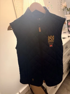 Marinblå quiltad dunväst Morris - Snygg marinblå dunväst från Morris med quiltat mönster och guldfärgad brodyr på bröstet samt brittisk flagga. Västen har hög krage och dragkedja framtill med läderdetalj. Perfekt för lager-på-lager och stilren look. Storleken är xsmall. Men kan med 100% säga att den passar folk som är s. Jag är 176 och den passar mig.