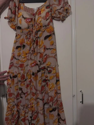 Blommig långklänning med puffärm - Långklänning med blommigt mönster i gult, orange och rött på beige botten. Klänningen har puffärmar, knytning framtill och volangdetaljer. Perfekt för sommarens varma dagar och har en romantisk vibe.