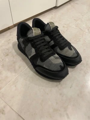 Valentino Garavani camo sneakers - Snygga sneakers från Valentino Garavani med grått och svart camo-mönster. Skorna har svarta detaljer, mesh- och mockapartier samt klassisk låg siluett. Perfekta för dig som vill sticka ut med stilrena och trendiga sneakers.