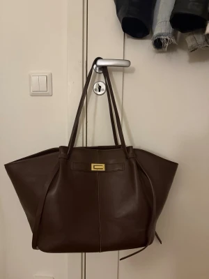 Brun shoppingväska i läder - Stilren brun shoppingväska i slätt läder med långa axelband och dekorativt spänne i guld framtill. Väskan har en rymlig design med utstickande sidor och enkel, modern look. Perfekt för dig som vill ha plats med allt du behöver. Ena bandet är sönder men går att limma ihop (det är inte helt av så går ändå att använda väskan som den är nu