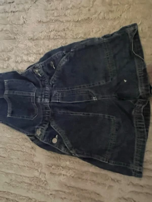 Mörkblå jeanskjol med hängslen - Säljer en mörkblå jeanskjol med hängslen och stora fickor framtill. Kjolen har knappar i sidorna och klassiska jeanssömmar. Perfekt för dig som gillar en avslappnad och trendig stil.