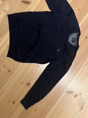 Mörkblå stickad V-neck tröja från Gant - Snygg mörkblå stickad tröja från Gant med liten broderad logga på bröstet. Tröjan har rund halsringning och långa ärmar. Perfekt till jeans eller chinos för en clean och klassisk look.