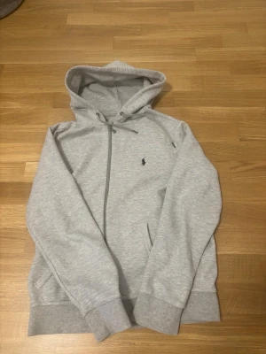 Ljusgrå hoodie från Polo Ralph Lauren - Ljusgrå hoodie med dragkedja från Polo Ralph Lauren. Klassisk design med huva och dragsko, två fickor framtill och broderad logga på bröstet. Tillverkad i mjukt bomullsmaterial för en skön och avslappnad stil.