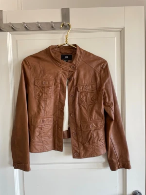 Brun skinnjacka från H&M - Snygg brun skinnjacka från H&M med fyra fickor framtill och knappar. Jackan har en rak passform, ståkrage med knapp och dekorativa sömmar. Perfekt för dig som vill ha en stilren och trendig look. Den är i super-skick! Jag har inte använt jackan ute, endast provat den🤎