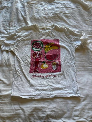 Vit t-shirt med färgglatt tryck - Vit t-shirt med färgglatt grafiskt tryck på framsidan som visar olika saker som bananer, dryck och texten 'Miscellany of things'. Klassisk passform och rund hals. Perfekt för dig som gillar unika motiv och en avslappnad stil.