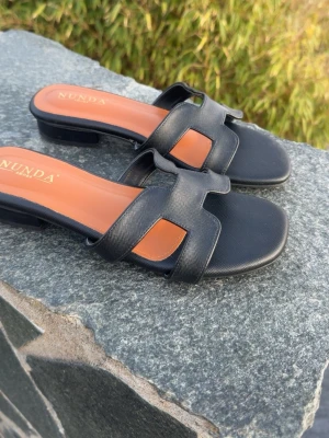 Svarta sandaler från NUNDA - Stilrena svarta sandaler från NUNDA med breda remmar i syntetmaterial och öppen tå. Sandalerna har en låg klack och en klassisk design med bruna innersulor. Perfekta för sommarens alla tillfällen.