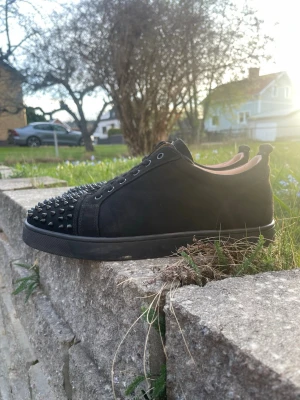 svarta Christian Louboutins med kvitto  - Säljer ett par svarta sneakers från Christian Louboutin med mattsvart mocka och coola nitar på tån. Skorna har klassisk låg profil, svarta skosnören och en svart sula. Perfekt för dig som vill sticka ut med en edgy stil.