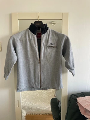 Grå ziptröja från Peak Performance - Snygg grå ziptröja från Peak Performance med hög svart krage och dragkedja framtill. Tröjan har en liten bröstficka med dragkedja och diskret logga på vänster sida. Perfekt för lager-på-lager och chill vardagsstil.