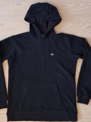 Lacoste hoodie  Storlek 16 (176cm) - Svart hoodie med Lacoste logga på bröstet och en ficka framtill. Storlek 16 som motsvarar 176 cm. Pris kan diskuteras.  Bra skick på tröjan.