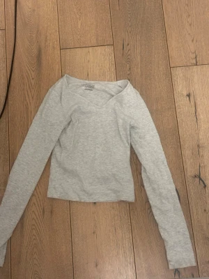 Ljusgrå långärmad topp från H&M - En enkel och stilren ljusgrå långärmad topp från H&M. Toppen har en v-ringad hals och är croppad, vilket ger en modern look. Perfekt att matcha med jeans för en avslappnad stil.