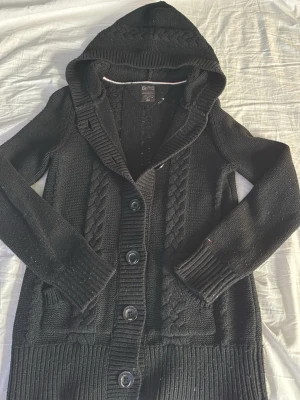 Stickad cardigan - Säljer en snygg mörkbrå stickad hoodie med flätade detaljer och knappar framtill. Perfekt för kyliga dagar med sin mysiga huva och långa ärmar. Ribbstickade muddar ger en skön passform. Passar både till jeans och mer avslappnade outfits. Storlek S men passar XS. Hör av er vid intresse!