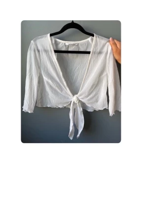 Vit transparent knytblus från Goldmann - Snygg vit transparent blus från Goldmann med knyt framtill och långa ärmar. Blusen har en djup v-ringning och är gjord i ett tunt, lätt material som ger en luftig känsla. Perfekt för lager på lager och trendiga outfits.