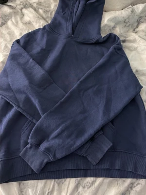 Mörkblå hoodie med huva - En enkel och stilren mörkblå hoodie med huva och magficka. Hoodien har ribbade muddar vid ärmslut och nederkant. Perfekt för en avslappnad och bekväm look. Materialet känns mjukt och skönt mot huden.