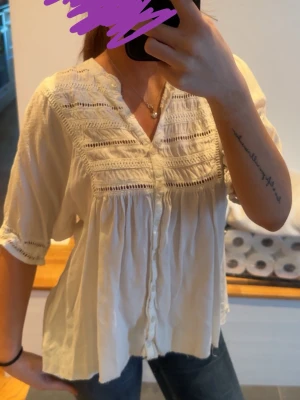 Vit boho-blus med spetsdetaljer - Söt vit blus i boho-stil med v-ringning och knappar framtill. Blusen har fina spetsdetaljer och broderade partier över bröstet samt luftiga, korta ärmar. Materialet ser ut att vara lätt och mjukt, perfekt för en avslappnad look.