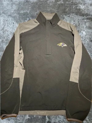 Baltimore Ravens sporttröja halvzip - Mörkgrå och beige långärmad sporttröja med halvzip och hög krage. Broderad Baltimore Ravens-logga på bröstet. Tillverkad i ett lätt och funktionellt material, perfekt för träning eller chill. Snygg kontrast mellan färgerna och sportig look.