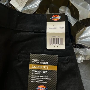 Svarta Dickies Twill Work Pants Loose Fit - Svarta arbetsbyxor från Dickies i twill-material. Loose fit med raka ben och flera fickor, inklusive en praktisk mobilficka. Klassisk Dickies-logga på bakfickan och benet. Perfekta för dig som gillar en avslappnad och rymlig passform. Helt nya, hann aldrig skicka tillbaka då de var för små för mig.