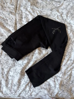 Y2K leggings med strass pistoler - Säljer ett par svarta kostym leggings med snygga strassdetaljer i form av pistoler vid benen. De har hög midja och är stretchiga för en bekväm passform. Perfekta till en avslappnad outfit eller när du vill ha något enkelt men ändå med lite extra bling. PERFEKT ATT HA UNDER STÖVLAR!!!! RIKTIGA 2000s Y2K fick från min faster men är tyvärr inte min stil💗 postar endast helger!!!!
