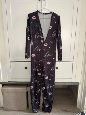 Svart bodypyjamas med rosa tryck och djup V-ringning - Säljer en svart långärmad pyjamas med djup V-ringning och knappar framtill. Pyjamasen har rosa tryck med läppar, hjärtan, läppstift och texten 'love' över hela plagget. Mjukt och stretchigt material som sitter skönt på kroppen.