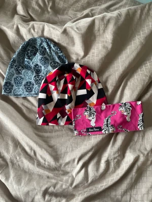 Två Anna Panna mössor o Ett Anna Panna pannband - Säljer ett set med två mössor och ett pannband. En mössa är blå med döskallemönster, den andra har ett geometriskt mönster i rött, svart, beige och vitt. Pannbandet är rosa med tigrar och har Anna panna-märke. Alla är i mjukt material, den bli har mindre passform (typ för barn) men de andra två passar bra för vuxna. Går att köpa separat. (Mössa: 100:- Pannband: 50)